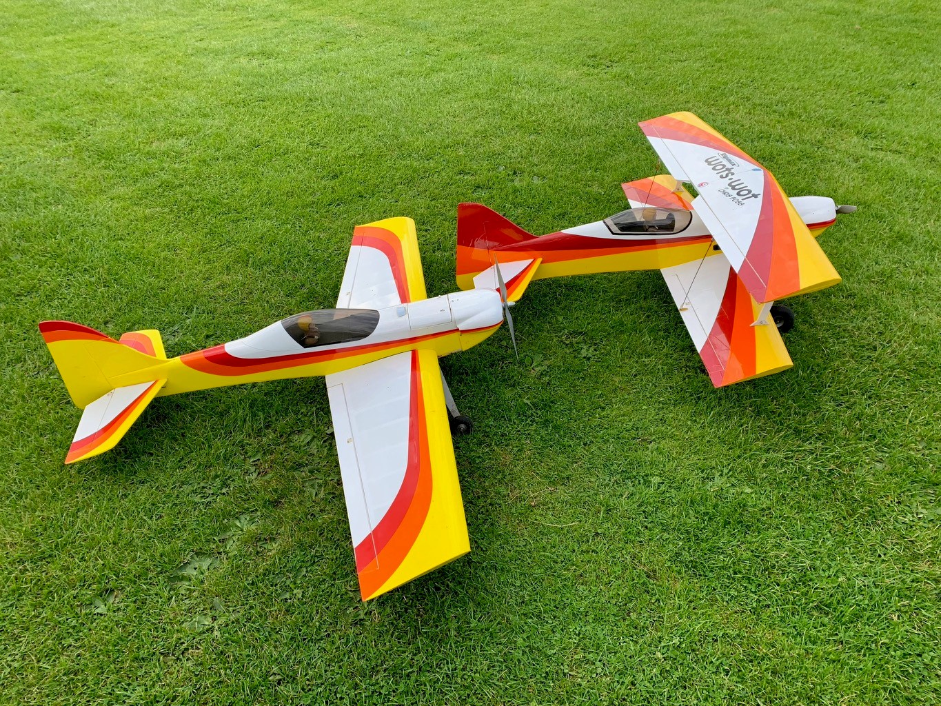 Voorkant -Tamiya || Kyosho Verkoopwinkel Two brightly coloured model aircraft in a field